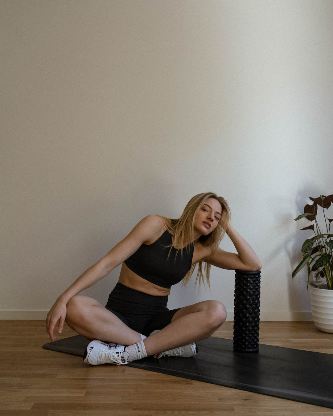 Blonde woman in sports bra and biker shorts sitting on a yoga mat leaning on a foam roller